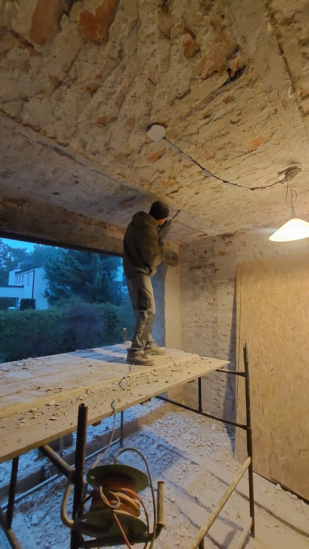 Pracownik na rusztowaniu, w trakcie skuwania tynku ze ściany z cegły wewnątrz budynku, widoczne fragmenty instalacji elektrycznej na suficie.