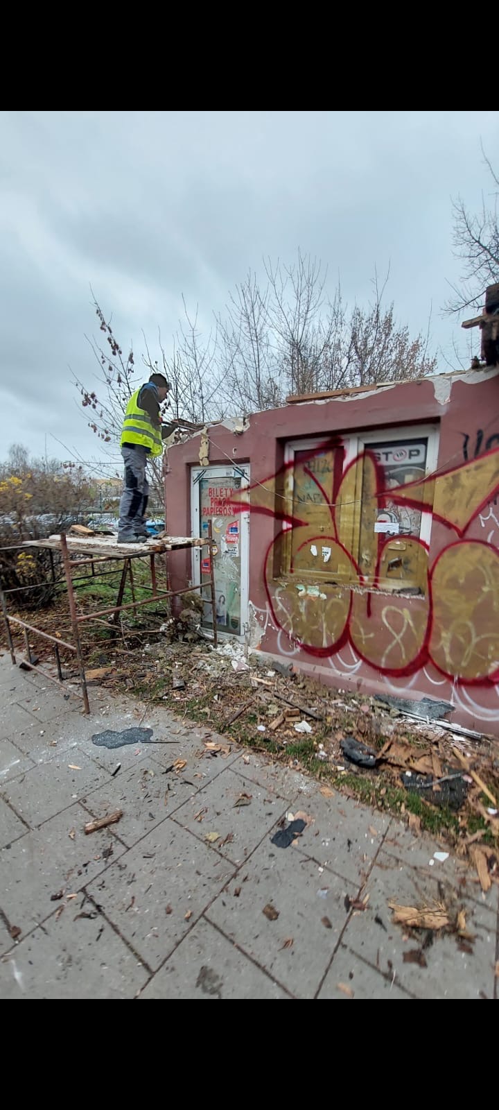 Pracownik w kamizelce odblaskowej na rusztowaniu demontuje fragment budynku z pomalowaną graffiti ścianą, widoczne ślady po wyburzeniach i gruz.
