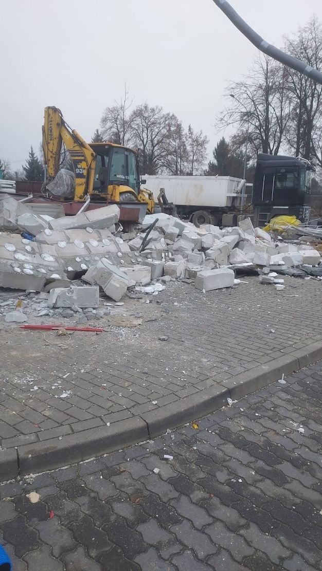 Żółta koparka i gruzowisko betonowych bloków na kostce brukowej, w tle ciężarówka na tle pochmurnego nieba i drzew.
