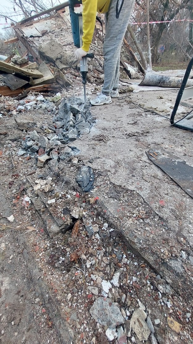 Pracownik w żółtej bluzie i białych rękawicach używa młota pneumatycznego do kruszenia betonowej posadzki na tle częściowo zburzonego budynku.