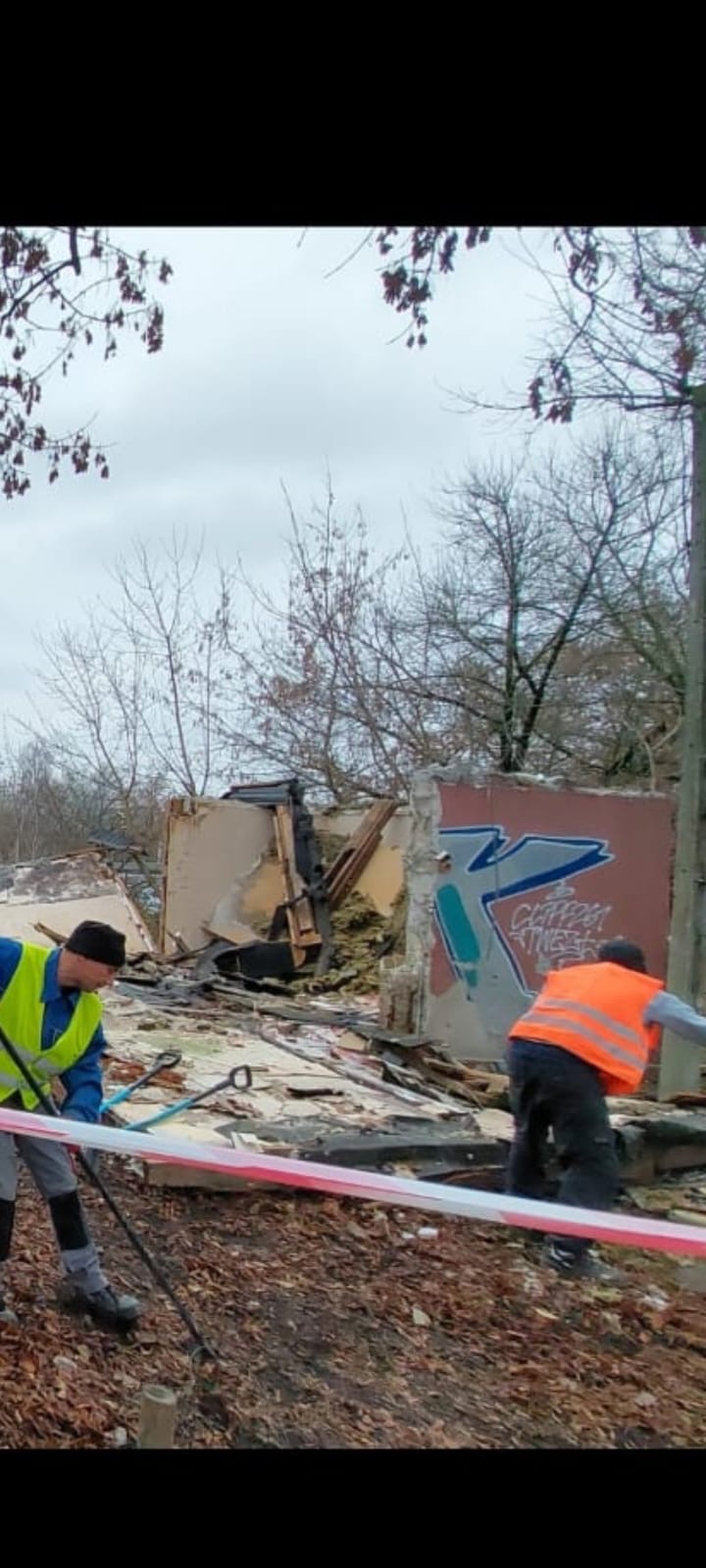 Dwóch pracowników w kamizelkach odblaskowych usuwa gruz z rozbiórki budynku, widoczne fragmenty ścian z graffiti i ostrzegawcza taśma.