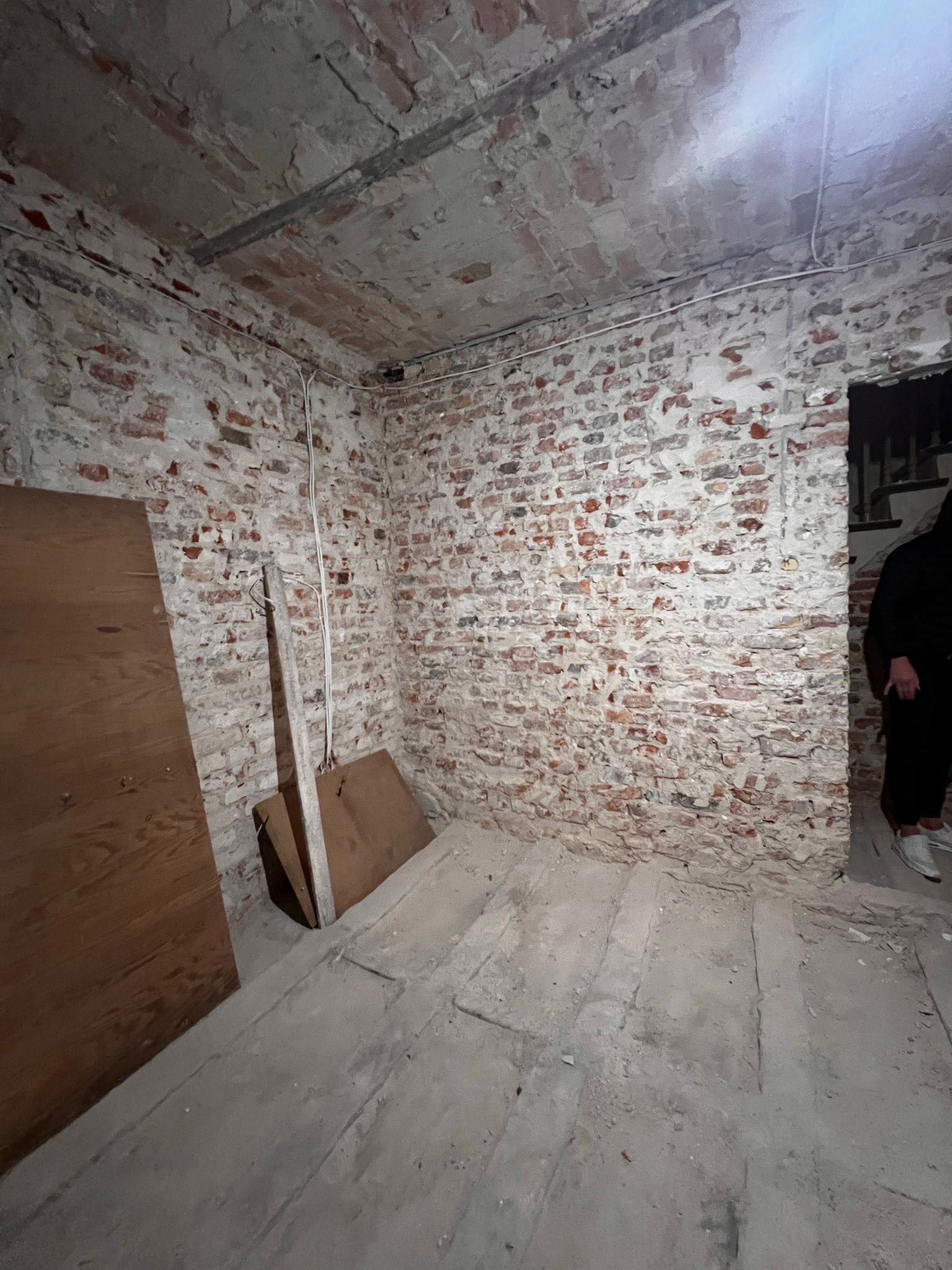 Wnętrze pomieszczenia z odsłoniętą, częściowo otynkowaną cegłą na ścianach, z widocznymi przewodami elektrycznymi, drewnianymi płytami opartymi o ścianę i osobą w drzwiach w tle.