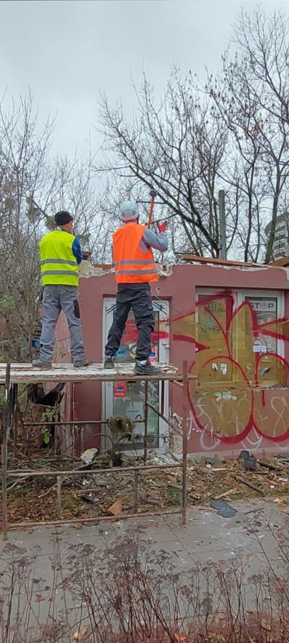 Dwóch pracowników w kamizelkach ochronnych na rusztowaniu, jeden uderza młotem w ścianę, budynek z graffiti w tle, drzewa bez liści.