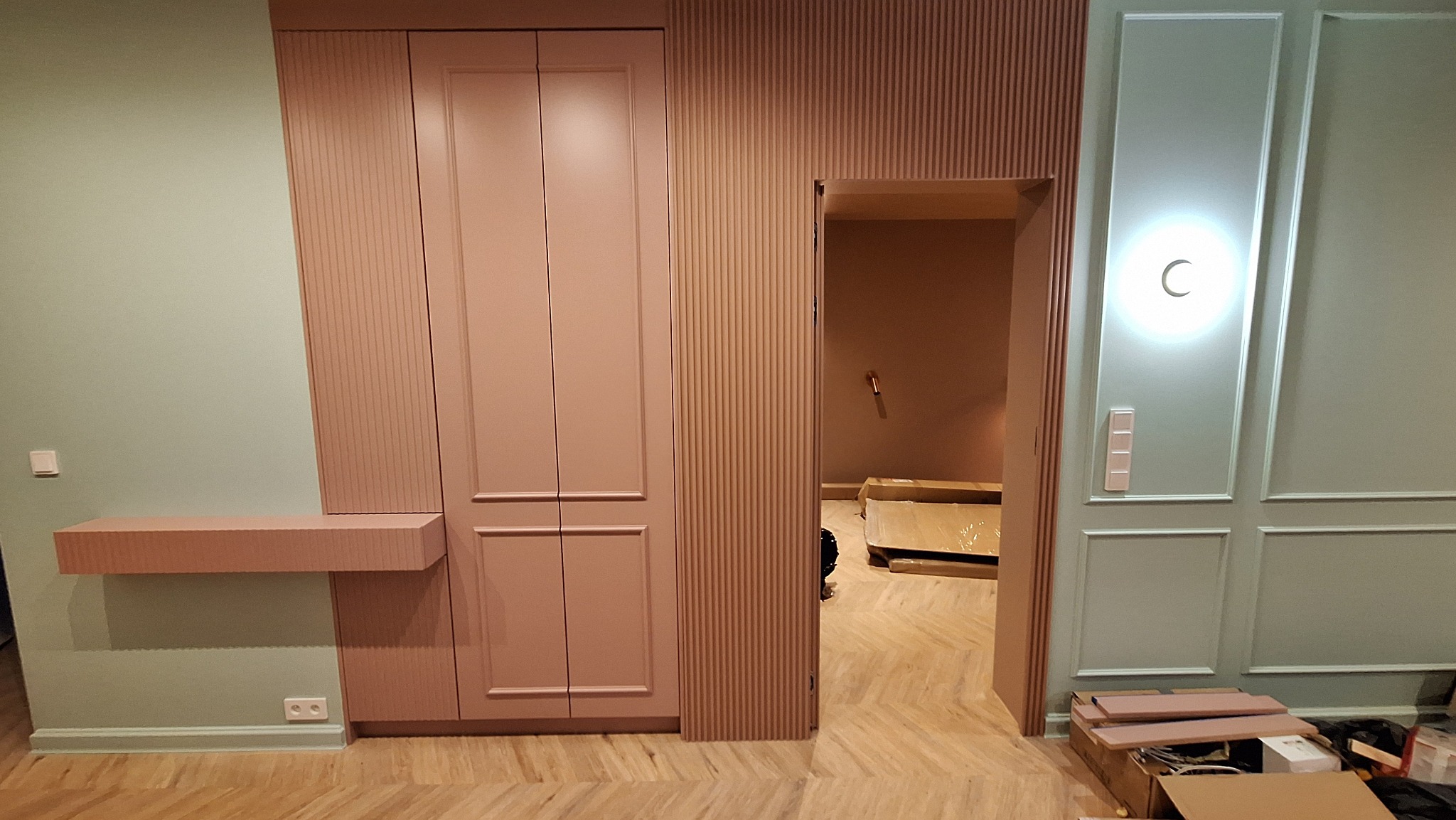 Pastelowe wnętrze z zabudową meblową: szafa w odcieniach różu z ryflowanymi panelami, półka i dekoracyjne listwy ścienne w kolorze miętowym.