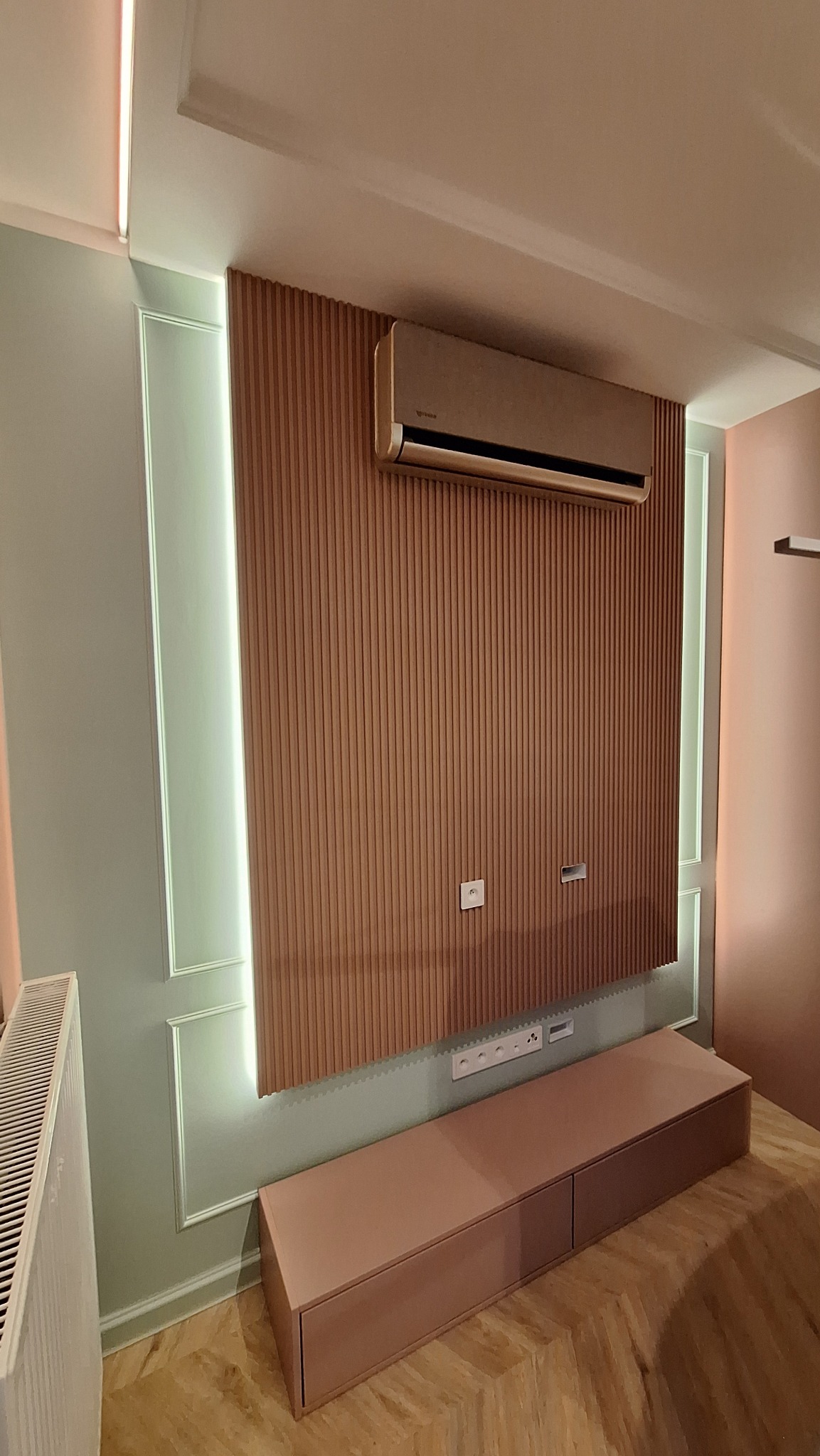 Nowoczesny salon: lamelowa ściana z klimatyzacją i podświetleniem LED, pastelowa komoda. Widoczny fragment grzejnika i parkiet w jodełkę. Minimalistyczny design.