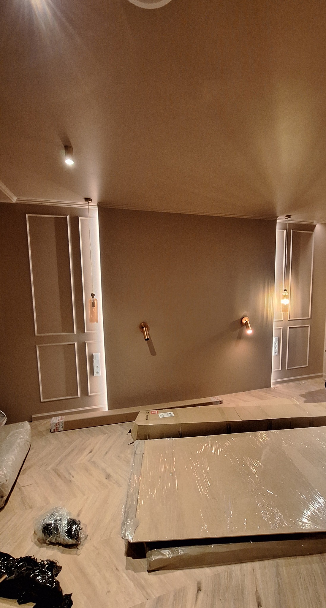 Eleganckie, beżowe wnętrze z ozdobnymi panelami ściennymi i nowoczesnym oświetleniem. Na podłodze panele ułożone w jodełkę, częściowo zakryte folią i kartonami.