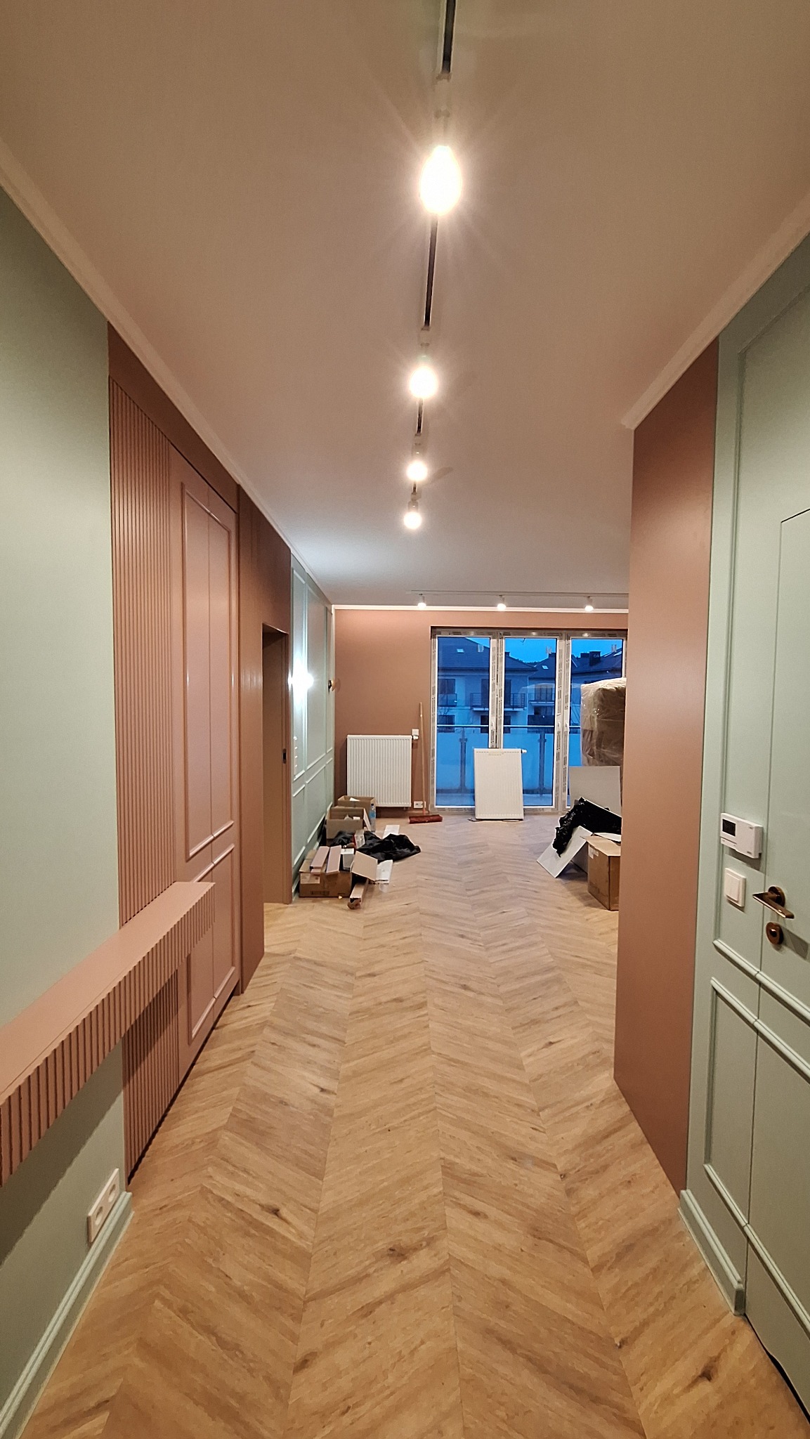 Wykończone wnętrze mieszkania z parkietem w jodełkę, pomalowanymi ścianami w odcieniach brązu i zieleni oraz nowoczesnym oświetleniem szynowym.