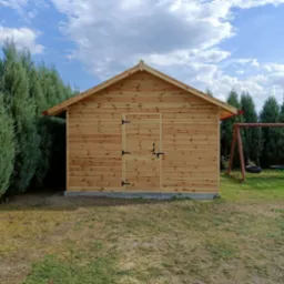 Mała, drewniana budowla gospodarcza z dwuskrzydłowymi drzwiami i czarnymi okuciami, otoczona zielonymi tujami i trawnikiem, z huśtawką w tle.