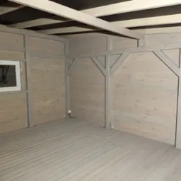 Wnętrze drewnianej konstrukcji w trakcie budowy lub wykańczania, widoczne drewniane ściany, podłoga z desek, okno z PCV oraz elementy konstrukcyjne dachu. Całość w jasnej, szarej tonacji.