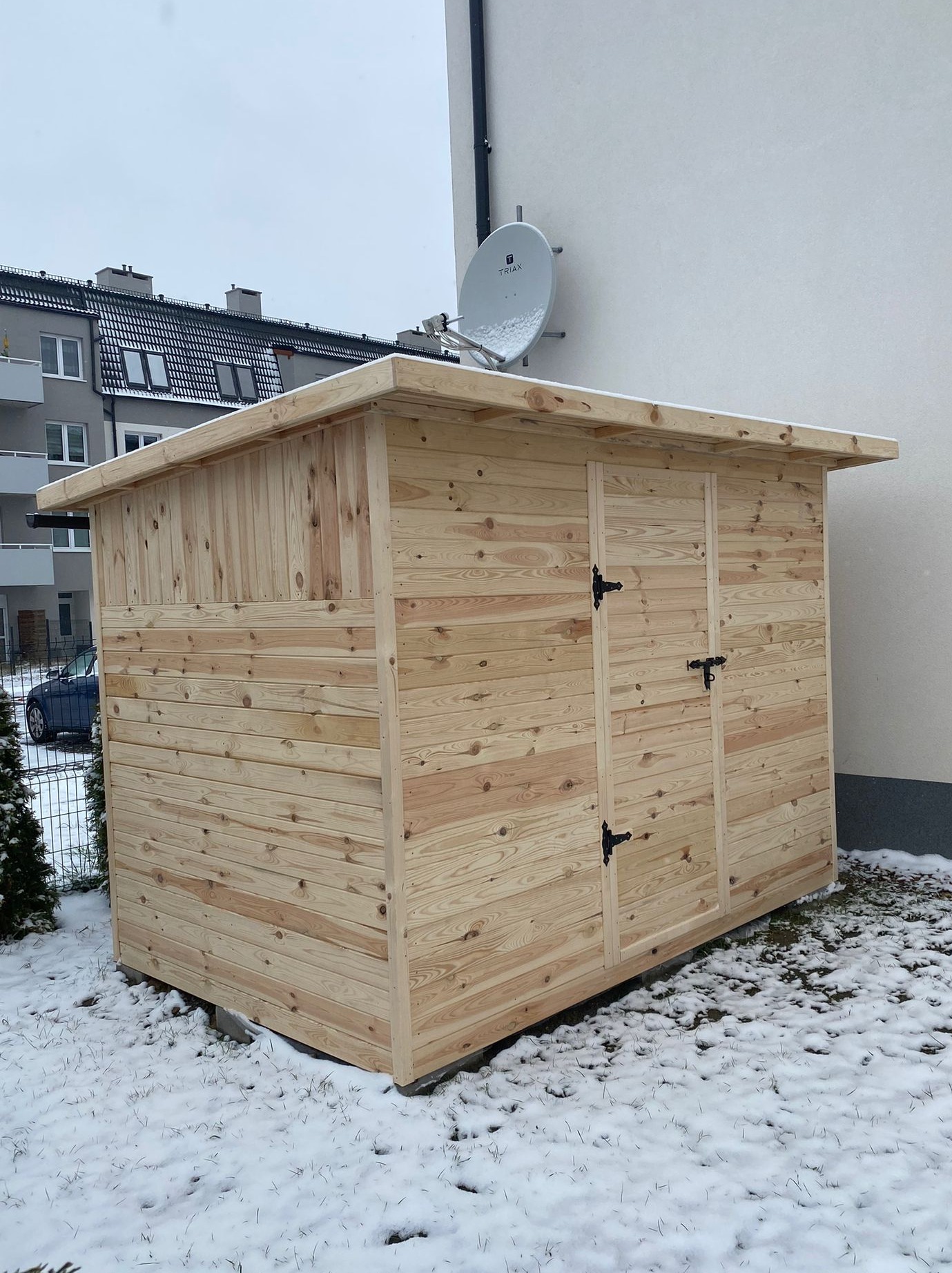 Drewniana, dwudrzwiowa szopa narzędziowa z jasnego drewna, z czarnymi okuciami, stoi na ośnieżonym trawniku przed budynkiem mieszkalnym.