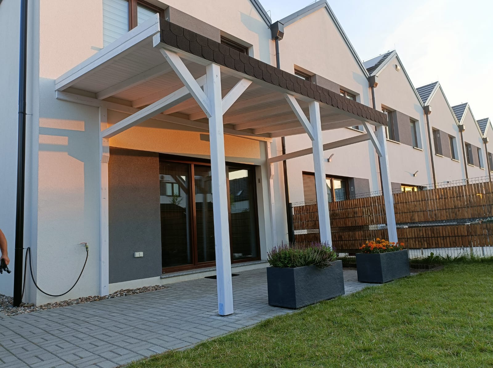 Biała pergola drewniana z brązowym pokryciem dachowym przylegająca do nowoczesnego domu szeregowego, z donicami z kwiatami na brukowanym tarasie.