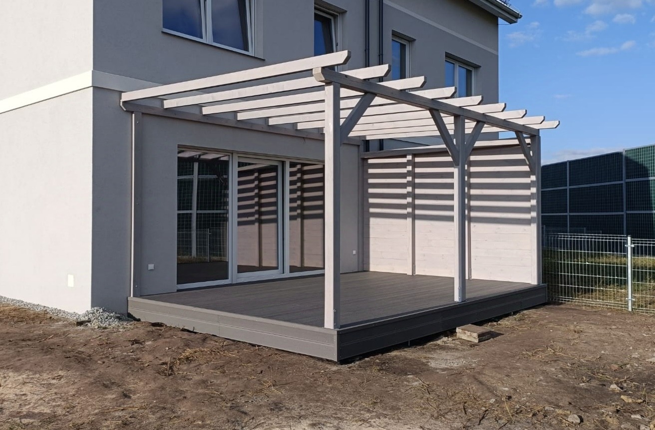Nowoczesna pergola z szarego drewna nad tarasem z desek kompozytowych, przylegająca do budynku z dużymi przesuwnymi drzwiami.