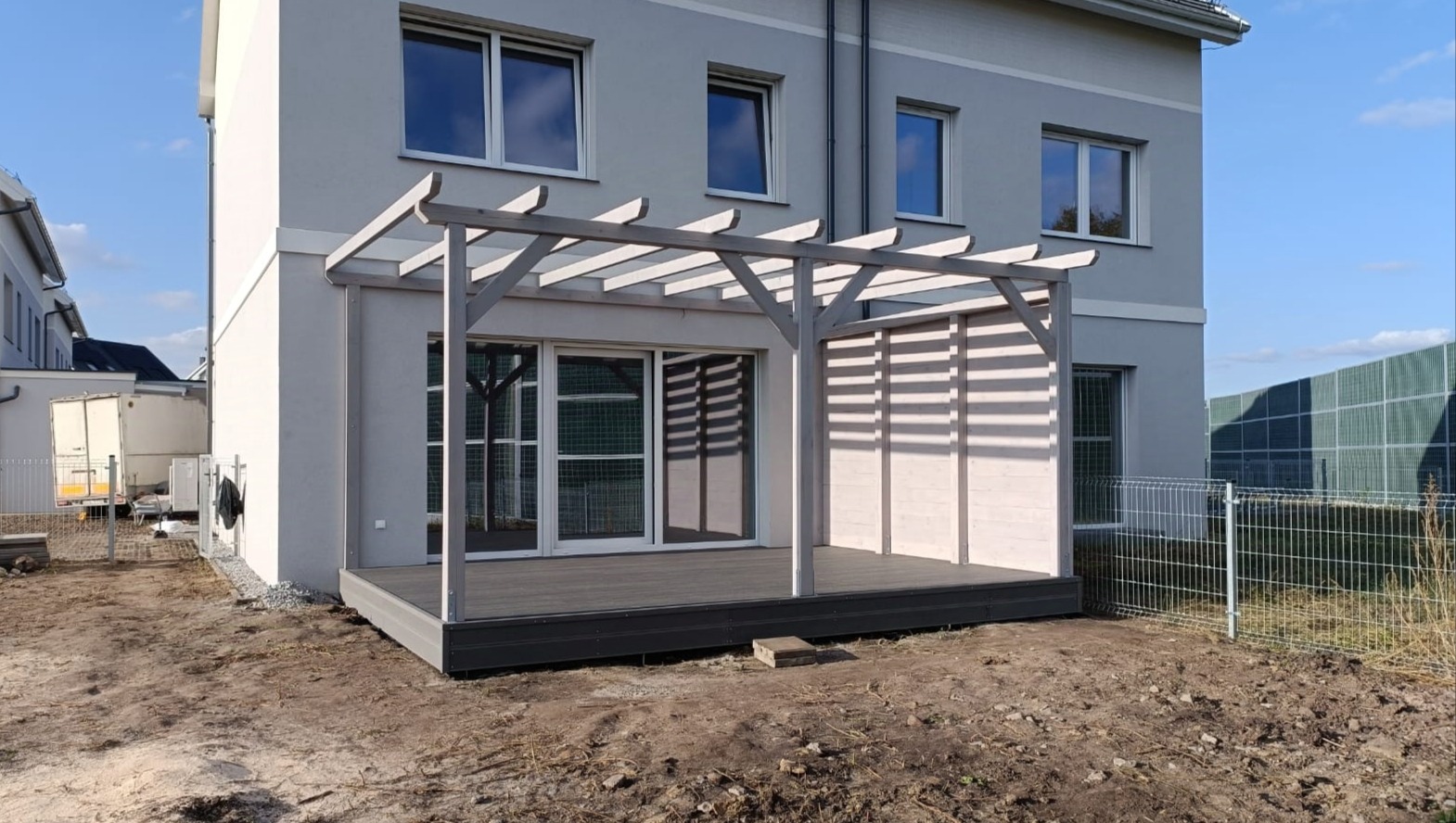 Szara pergola z drewnianą konstrukcją i tarasem przylegająca do nowoczesnego domu z dużymi oknami, teren wokół w trakcie prac ziemnych.