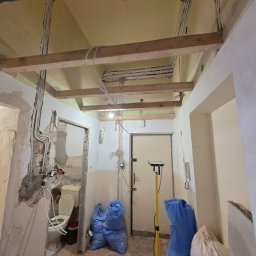 VoltSerwis Damian Bańdo - Wnętrze w trakcie remontu w Krakowie, z widocznymi przewodami elektrycznymi prowadzonymi pod sufitem, drewnianą konstrukcją, workami z materiałami budowlanymi i lampą pomiarową.