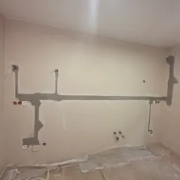 Ściana w trakcie prac elektrycznych: wyfrezowane bruzdy pod kable z widocznymi przewodami, gniazdkami i puszkami, częściowo zamaskowane zaprawą gipsową, podłoga zabezpieczona folią ochronną.