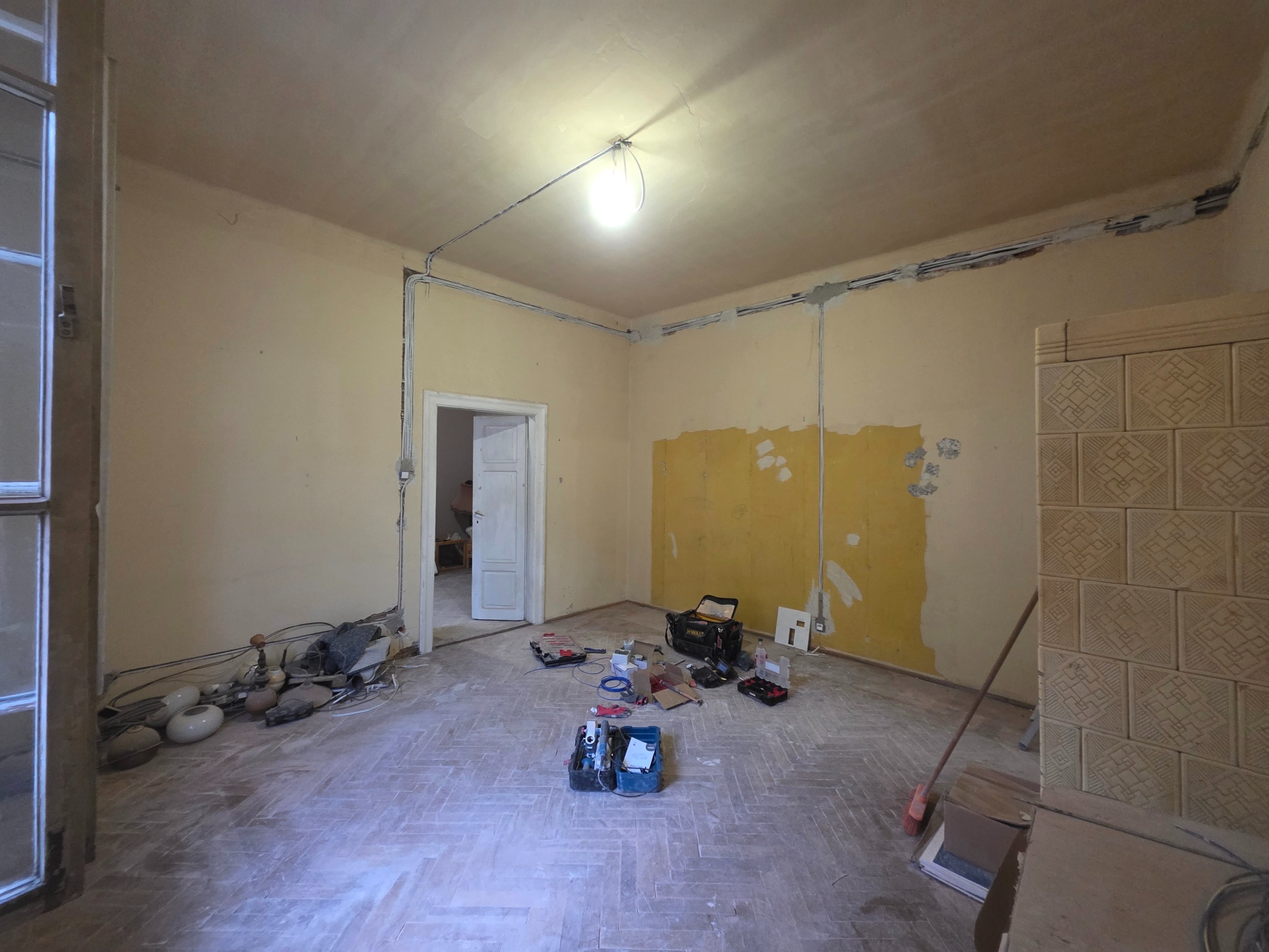 Pokój w trakcie remontu z widocznymi przewodami elektrycznymi prowadzonymi po ścianach i suficie, narzędzia rozłożone na podłodze, stara instalacja oświetleniowa z żarówką