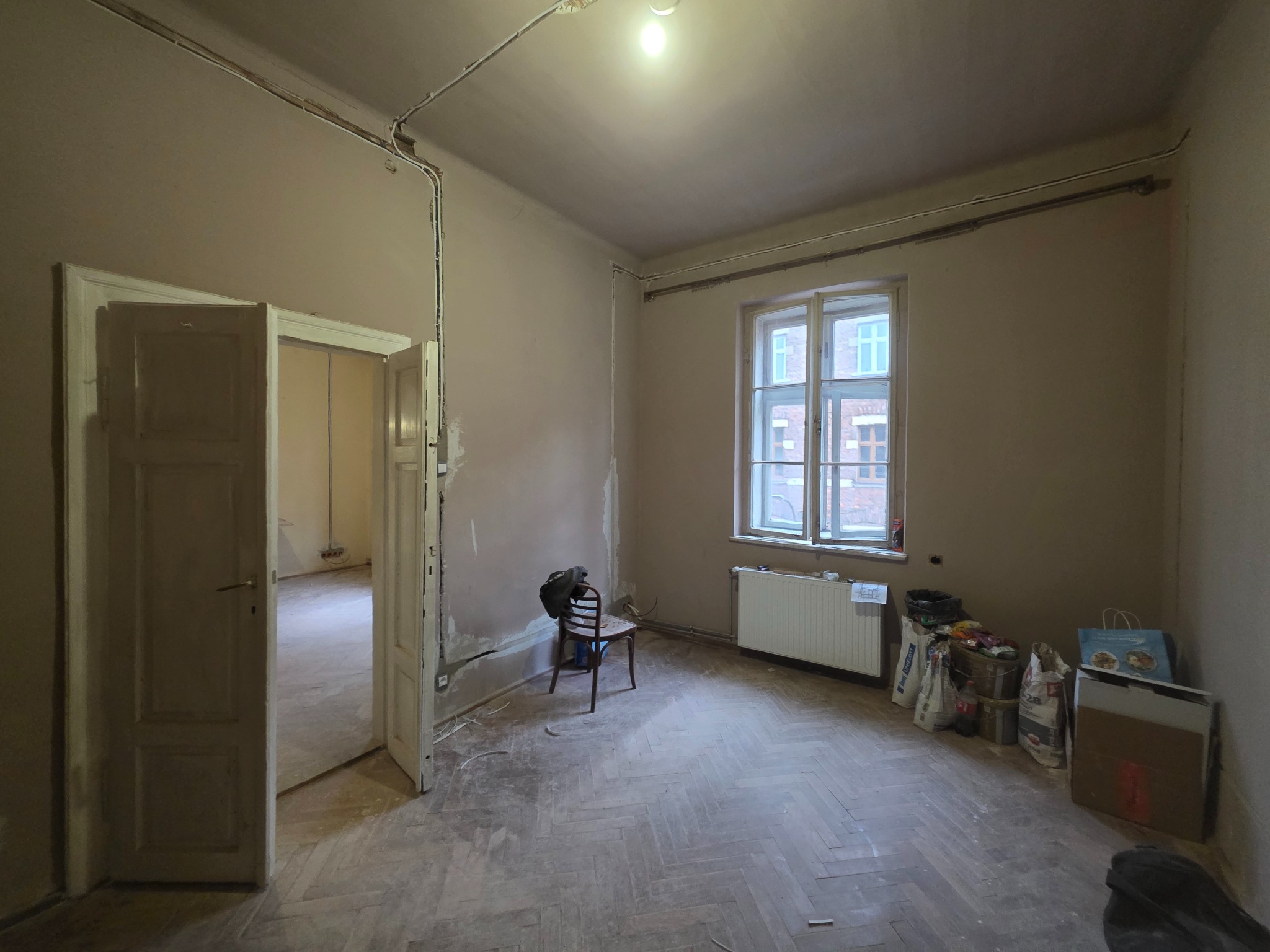 Pokój w trakcie remontu z widocznymi przewodami elektrycznymi na ścianie, otwartymi drzwiami, starym parkietem i workami z materiałami budowlanymi przy grzejniku.
