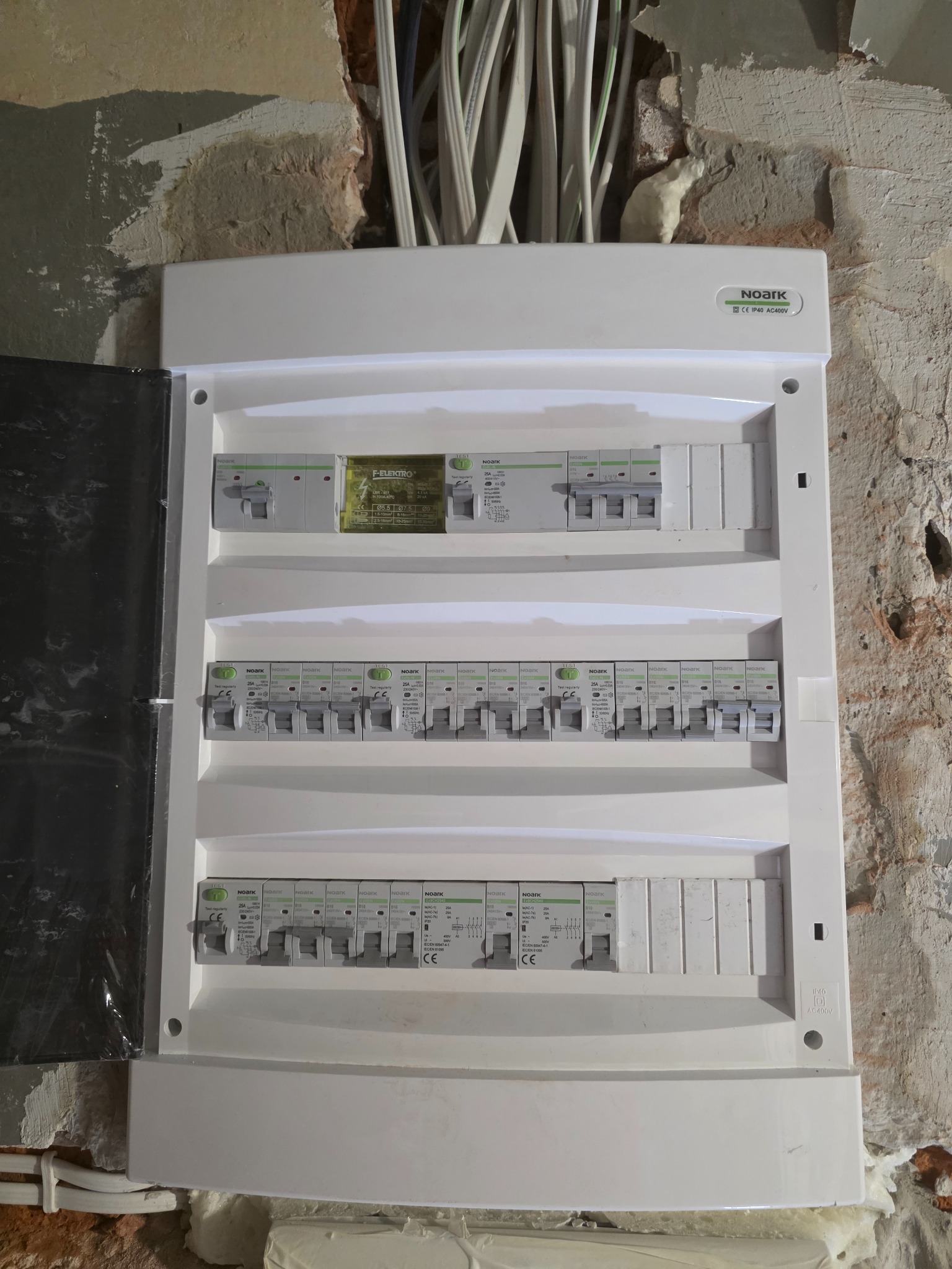 Rozdzielnica elektryczna Noark zamontowana na surowej, otynkowanej ścianie z widocznymi przewodami w górnej części, zabezpieczona pianką montażową w szczelinach.