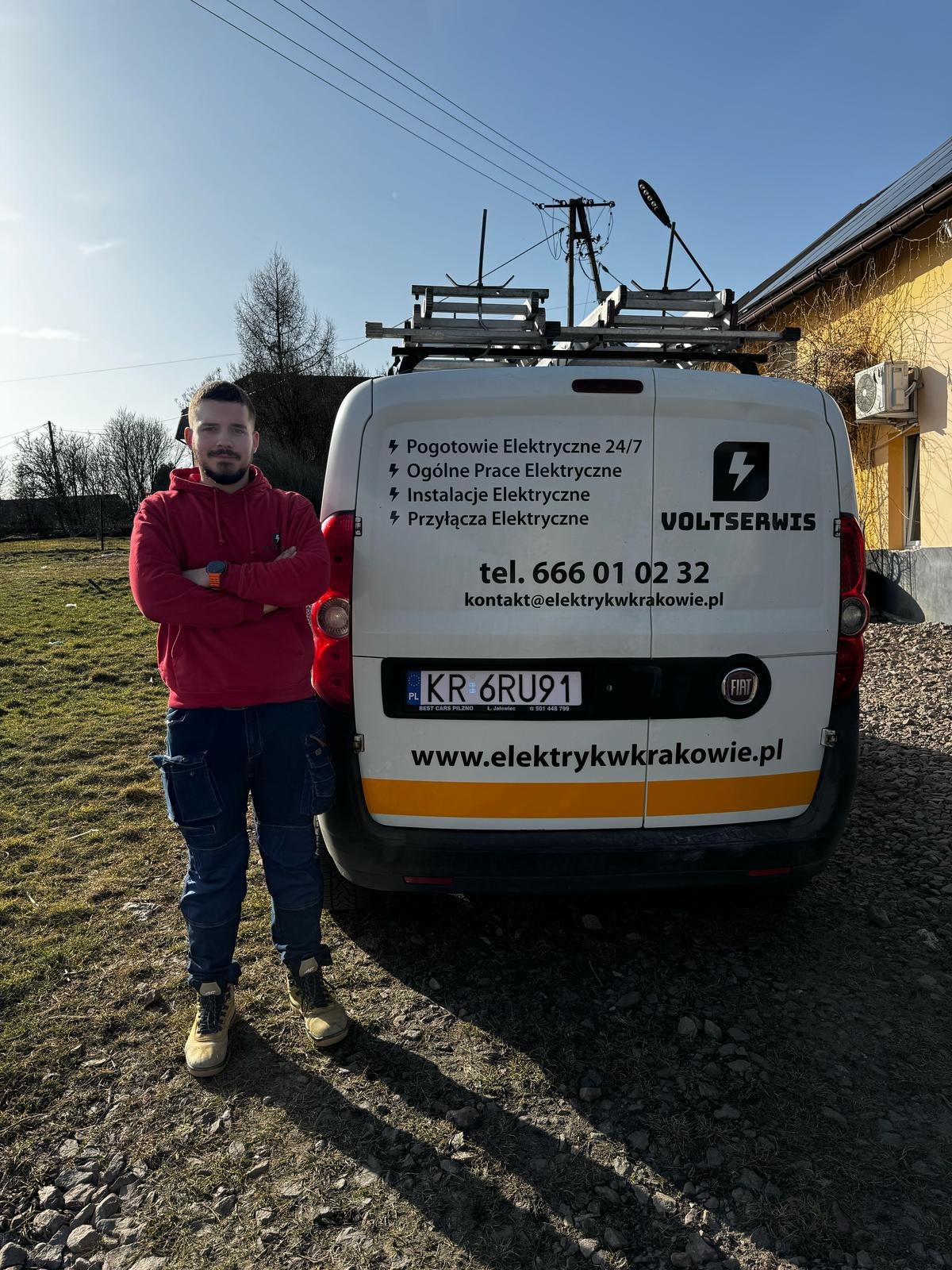 Elektryk w czerwonej bluzie stoi przed białym samochodem dostawczym firmy Voltservis z numerem telefonu i zakresem usług elektrycznych.