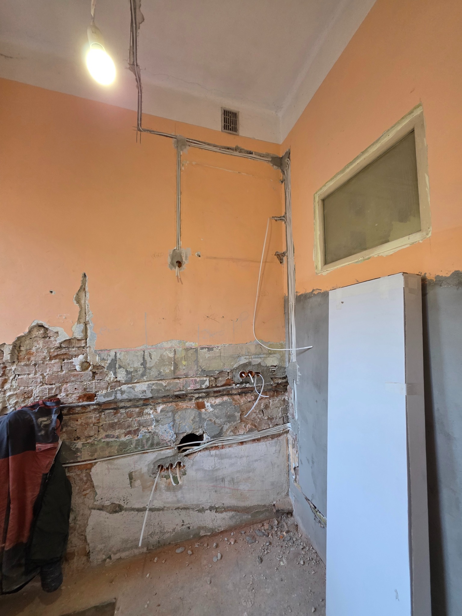 Wnętrze w trakcie remontu: widoczne przewody elektryczne w ścianie z odsłoniętą cegłą, świeżo położony tynk i fragment nowej szafy w rogu pomieszczenia.