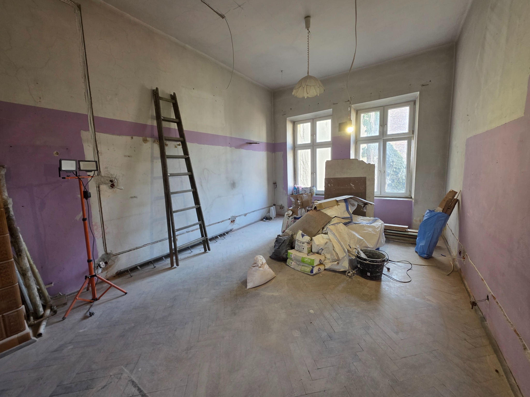 Pokój w trakcie remontu: widoczne ślady po instalacjach elektrycznych, drabina oparta o ścianę, worki z materiałami budowlanymi, lampa budowlana na trójnogu.