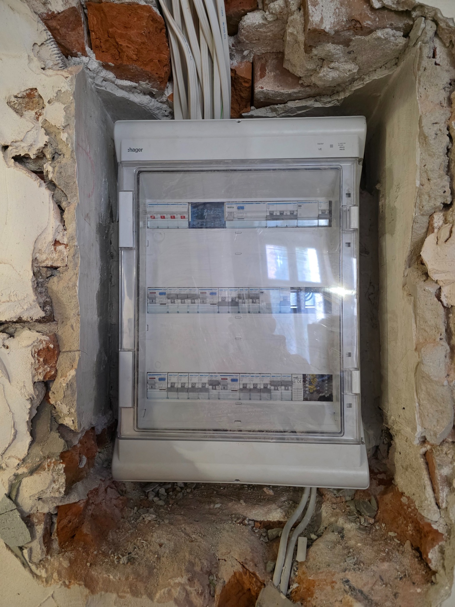 Rozdzielnica elektryczna Hager zamontowana w surowym, otynkowanym murze z widocznymi cegłami i przewodami elektrycznymi.