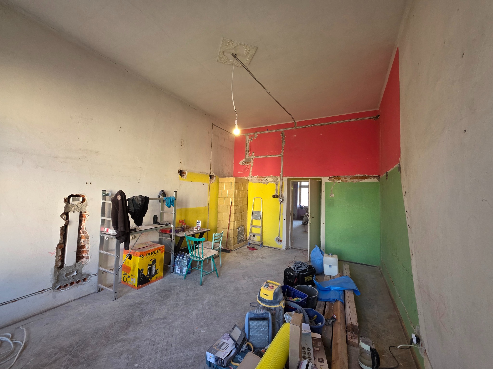Wnętrze remontowanego pokoju z widocznymi instalacjami elektrycznymi w ścianie, żółtym piecem kaflowym i narzędziami budowlanymi na podłodze.