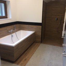 Adam Roszkowski - Nowoczesna łazienka z wanną obudowaną drewnem i kabiną prysznicową wykończoną płytkami imitującymi drewno. Szare płytki na podłodze i okno z drewnianą ramą.