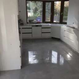 Jasna kuchnia w trakcie montażu: białe szafki dolne z czarnymi uchwytami, szare płytki podłogowe odbijające światło okna. Widok na zieleń za oknem.