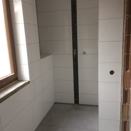 Adam Roszkowski - Wykończona łazienka z białymi płytkami, szarymi płytkami podłogowymi i pionowym pasem dekoracyjnym z ciemnych płytek w strefie prysznica. Widoczne okno z drewnianą ramą.