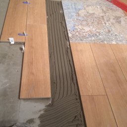 Adam Roszkowski - Układanie podłogi z płytek imitujących drewno, widoczna warstwa kleju, poziomica, krzyżyki dystansowe i fragment starej podłogi. Prace glazurnicze w toku.