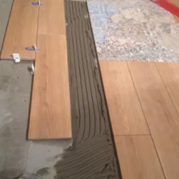 Układanie podłogi z płytek imitujących drewno, widoczna warstwa kleju, poziomica, krzyżyki dystansowe i fragment starej podłogi. Prace glazurnicze w toku.