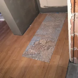 Ułożone panele podłogowe w odcieniach drewna z wstawką dekoracyjnej płytki w trakcie prac wykończeniowych, widoczne surowe ściany i narożnik.