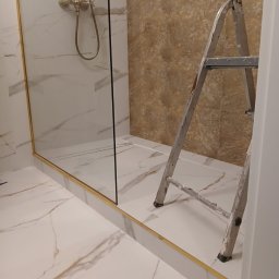 Adam Roszkowski - Wykończona kabina prysznicowa z marmurowymi płytkami i złotymi detalami. Widoczna drabina malarska, sugerująca trwające prace wykończeniowe w łazience.
