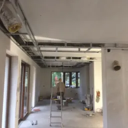 Sufit podwieszany z metalową konstrukcją w surowym stanie, widoczne przewody wentylacyjne. W tle osoba na drabinie, okno. Wnętrze w trakcie prac wykończeniowych.