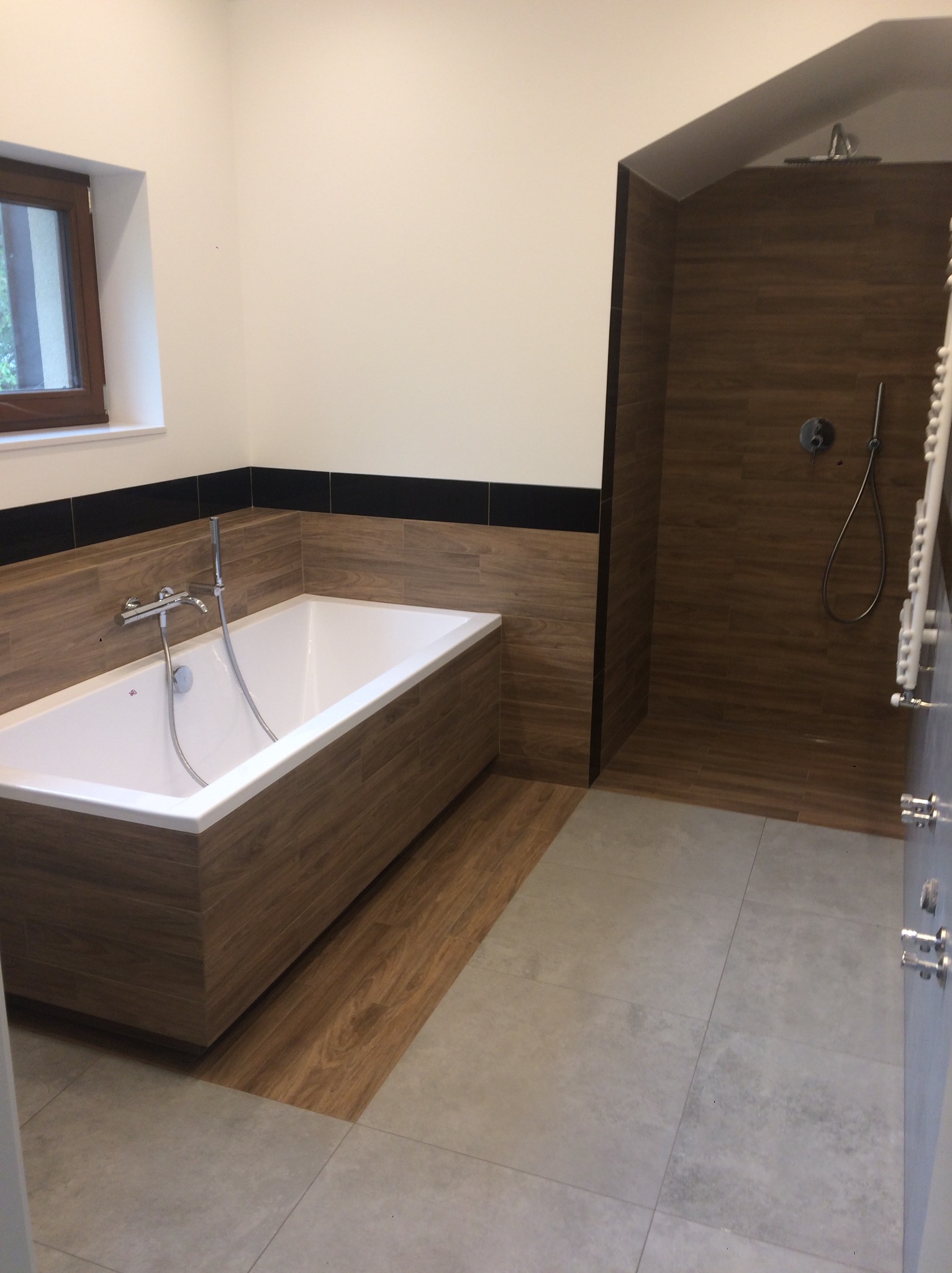 Nowoczesna łazienka z wanną obudowaną drewnem i kabiną prysznicową wykończoną płytkami imitującymi drewno. Szare płytki na podłodze i okno z drewnianą ramą.