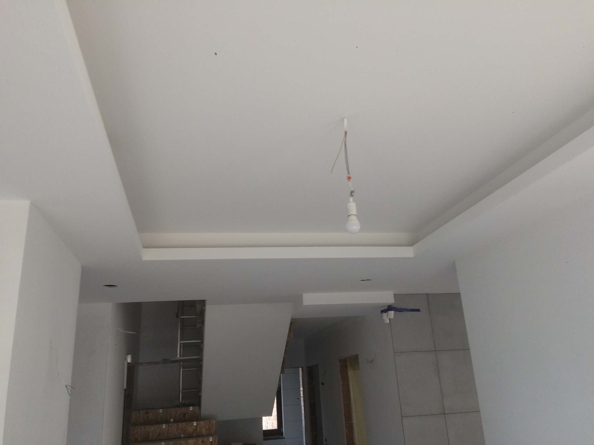 Biały sufit podwieszany z oświetleniem w trakcie wykańczania wnętrza. Widoczne kable i żarówka, w tle drabina i płytki na ścianie.