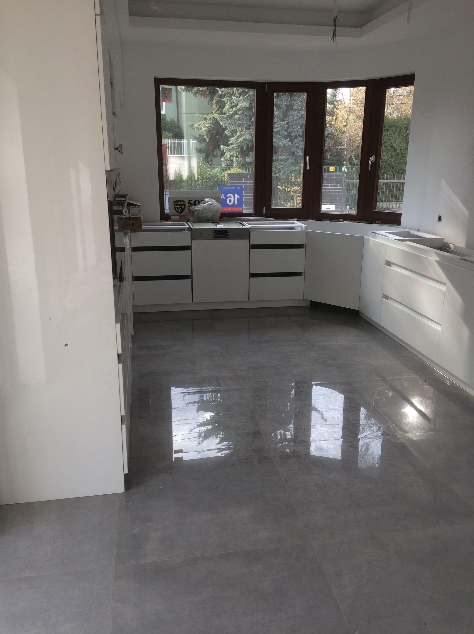 Jasna kuchnia w trakcie montażu: białe szafki dolne z czarnymi uchwytami, szare płytki podłogowe odbijające światło okna. Widok na zieleń za oknem.