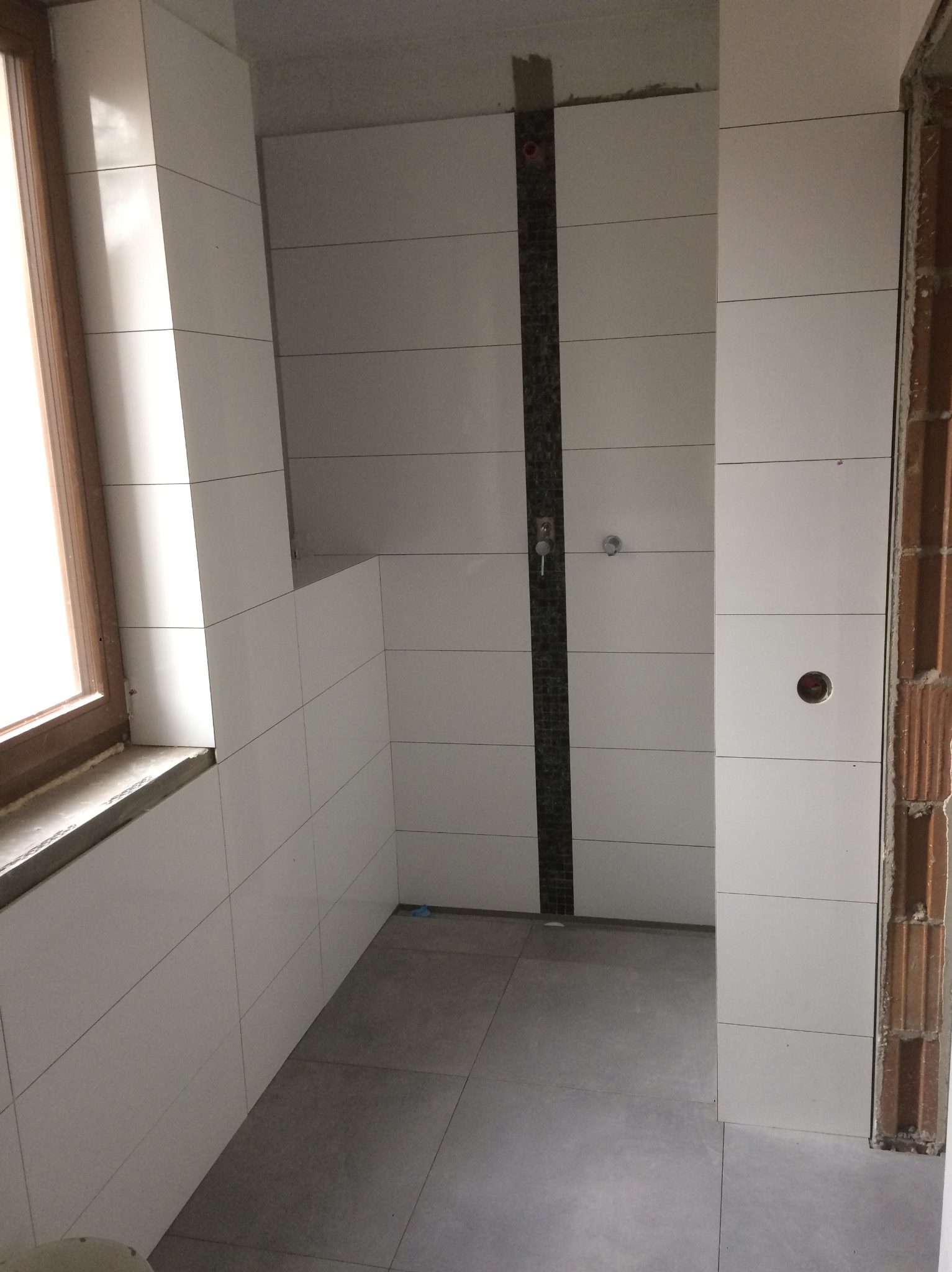 Wykończona łazienka z białymi płytkami, szarymi płytkami podłogowymi i pionowym pasem dekoracyjnym z ciemnych płytek w strefie prysznica. Widoczne okno z drewnianą ramą.