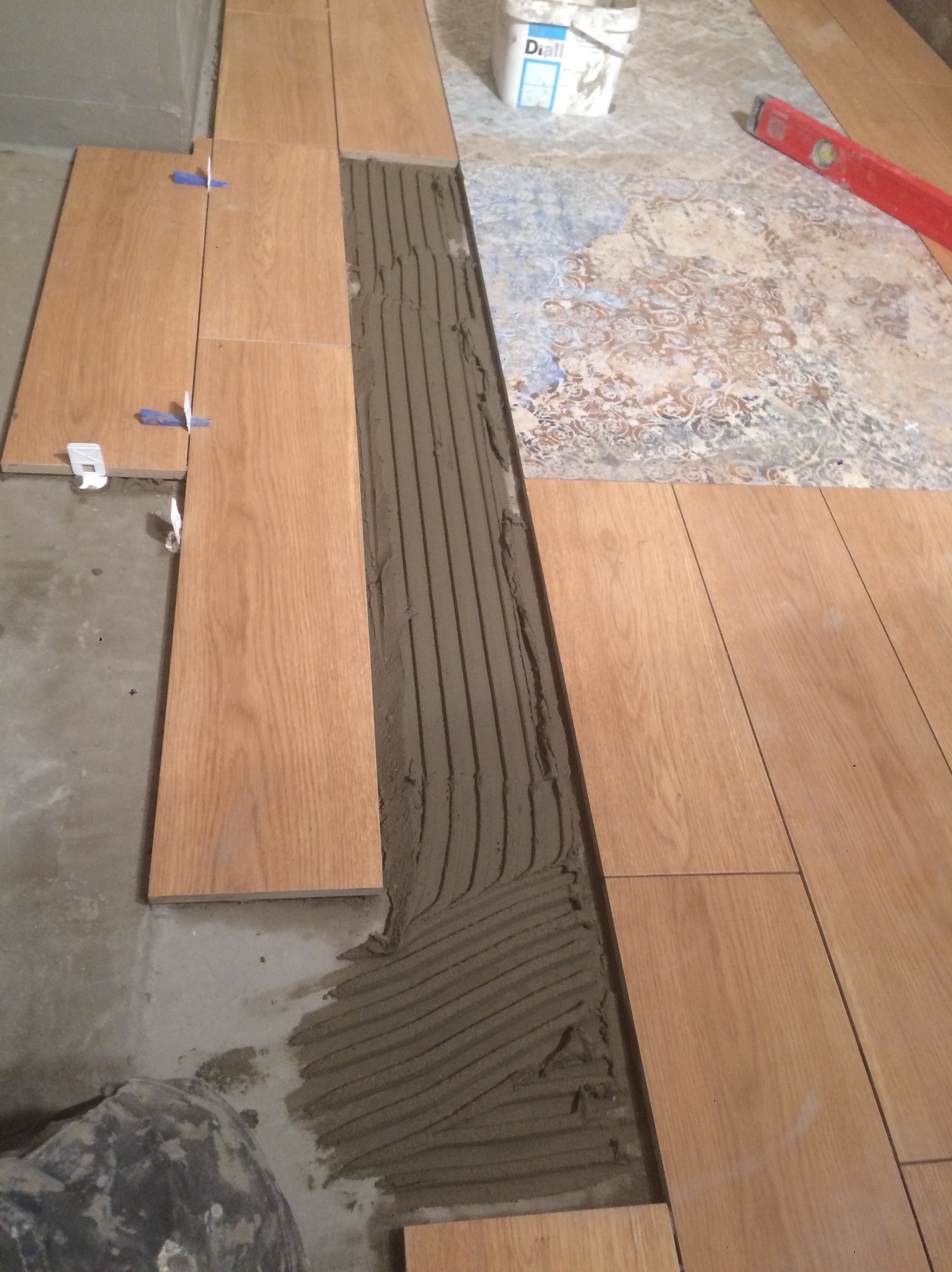 Układanie podłogi z płytek imitujących drewno, widoczna warstwa kleju, poziomica, krzyżyki dystansowe i fragment starej podłogi. Prace glazurnicze w toku.