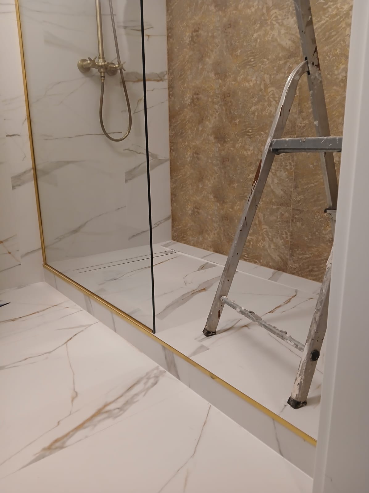 Wykończona kabina prysznicowa z marmurowymi płytkami i złotymi detalami. Widoczna drabina malarska, sugerująca trwające prace wykończeniowe w łazience.
