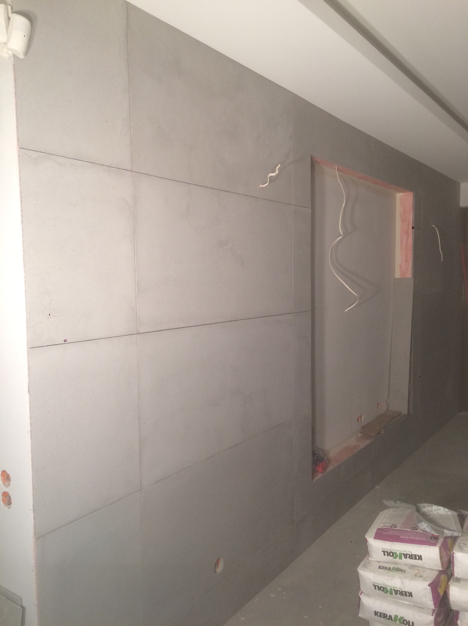 Minimalistyczna ściana wykończona szarymi płytami w dużej skali, z widocznym otworem na instalacje i workami z zaprawą na podłodze. Surowe wnętrze w trakcie prac wykończeniowych.