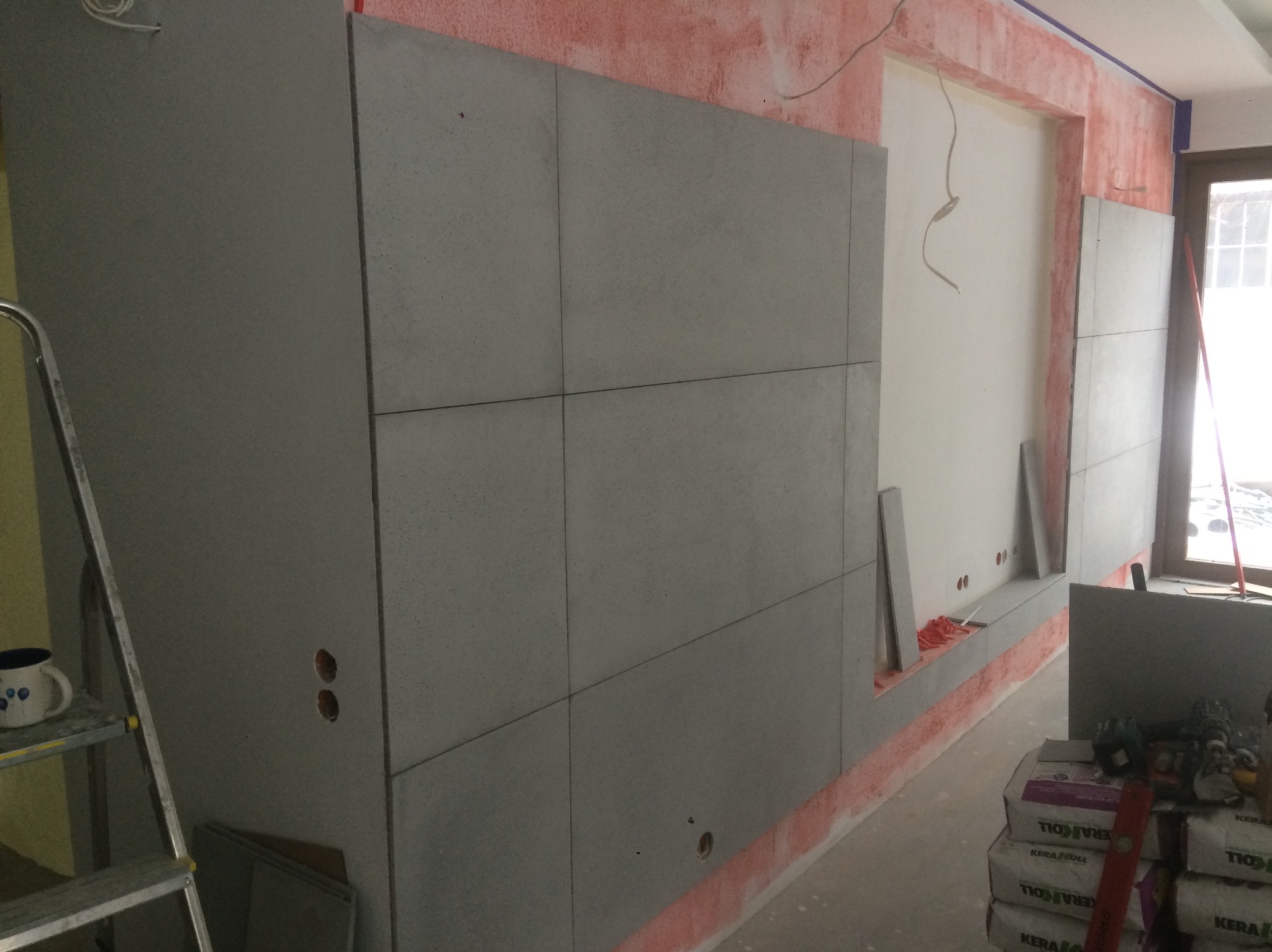 Montaż szarych płyt na ścianie z widocznymi otworami na gniazdka. Na podłodze leżą worki z klejem, a obok stoi drabina z kubkiem.