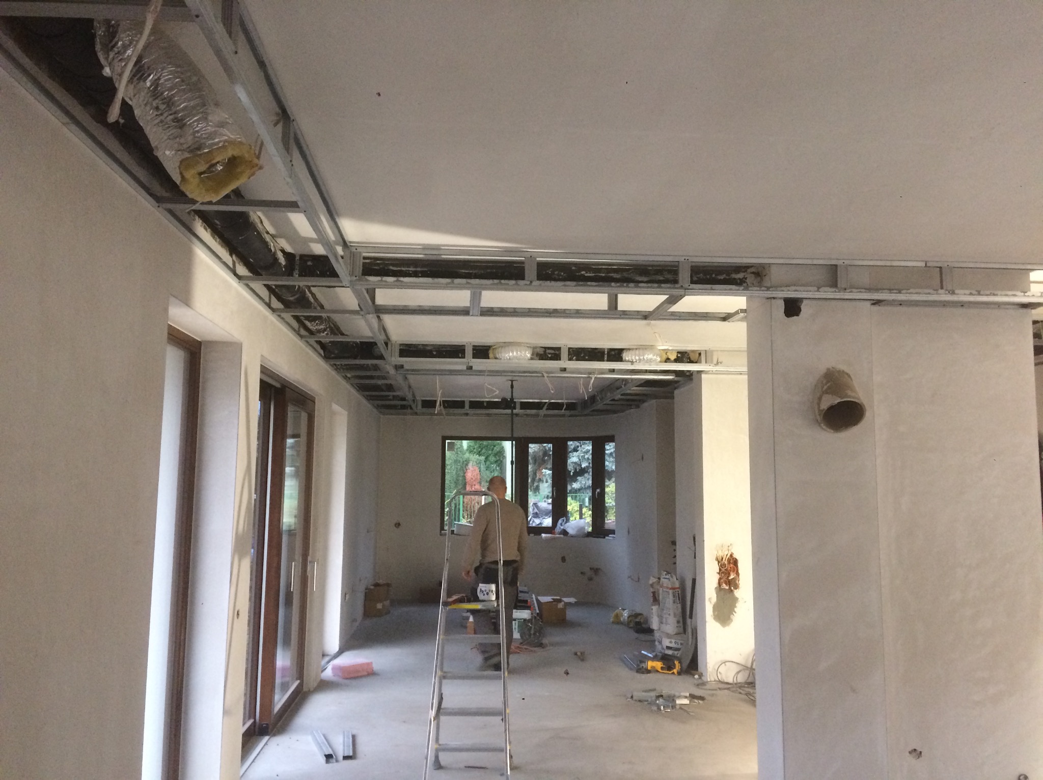 Sufit podwieszany z metalową konstrukcją w surowym stanie, widoczne przewody wentylacyjne. W tle osoba na drabinie, okno. Wnętrze w trakcie prac wykończeniowych.