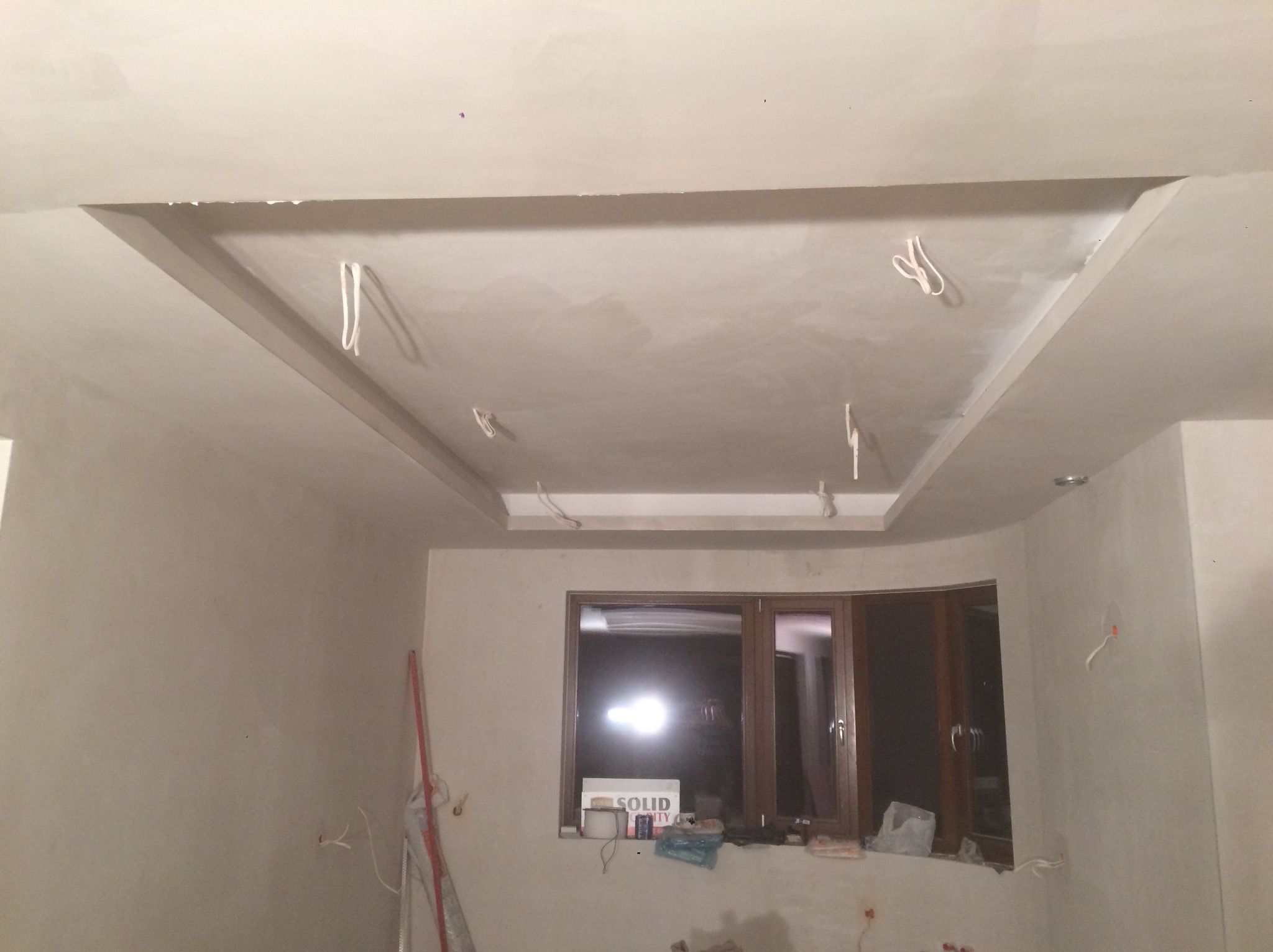 Wykończony sufit podwieszany z gładzią w jasnym wnętrzu. Widoczne kable oświetleniowe, ściany przygotowane do malowania. Okno z parapetem.