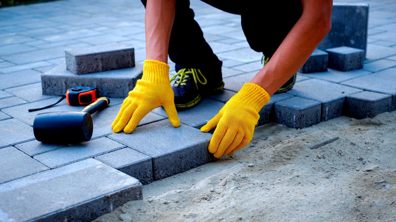 Układanie szarej kostki brukowej na piasku, widoczne żółte rękawice robocze, gumowy młotek i miara zwijana.