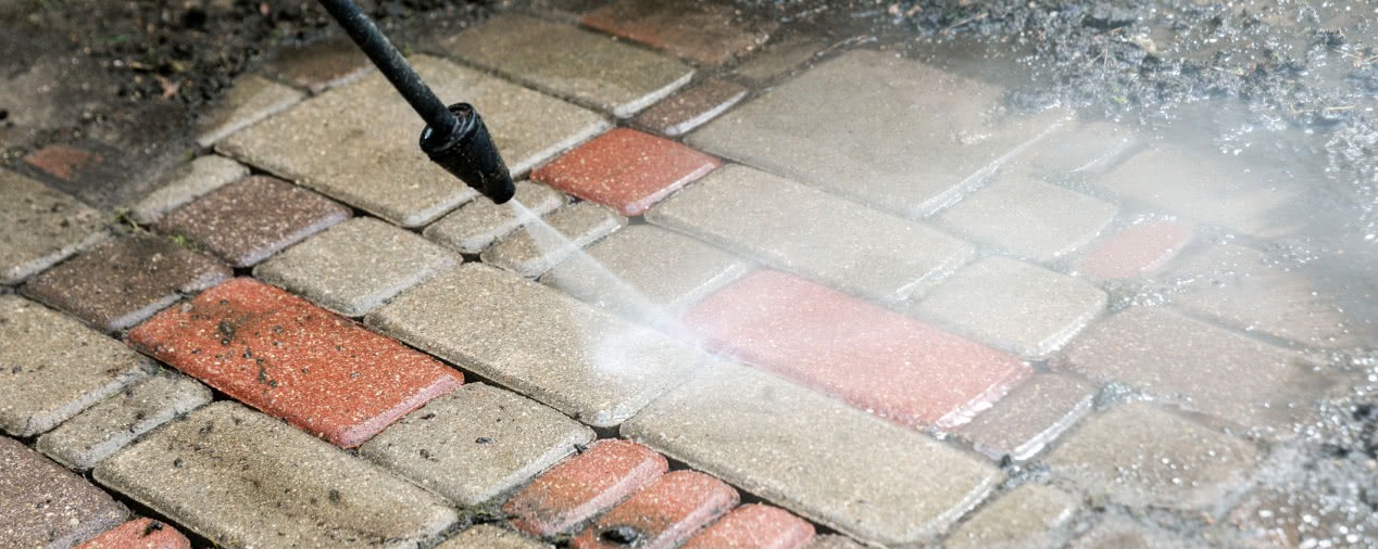 Czyszczenie kostki brukowej myjką ciśnieniową, woda spryskuje powierzchnię z bliska, widoczne zabrudzenia i różnica w kolorze po czyszczeniu.
