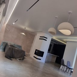 EL-PAT Instalacje elektryczne - Nowoczesne oświetlenie sufitowe w przestronnym salonie z kominkiem i szarymi płytkami podłogowymi. Widoczne lampy wiszące i listwy LED.