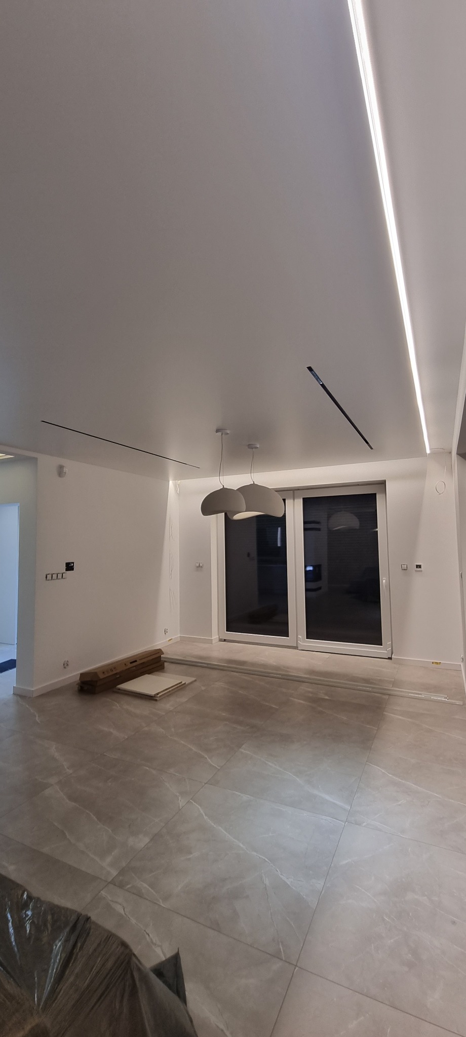 Nowoczesne wnętrze z oświetleniem LED w suficie i wiszącymi lampami. Widoczne duże okno balkonowe i płytki podłogowe. Częściowo widoczne materiały budowlane.