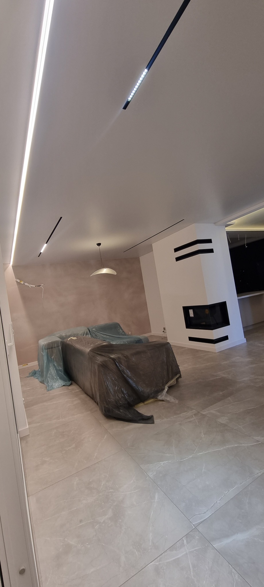 Nowoczesny salon w trakcie wykończenia: minimalistyczne oświetlenie LED, kominek z czarnymi akcentami, meble zabezpieczone folią ochronną, szare płytki podłogowe.